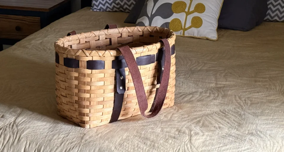 A handwoven reed tote basket with leather handles packed on a bed